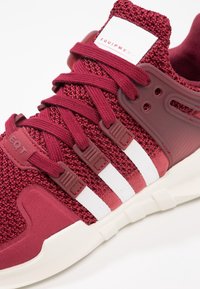 Bordové sportovní boty se strukturovaným pleteným svrškem, bílými pruhy, polstrovaným límcem a gumovou podrážkou. Má šněrovací design a syntetické detaily.