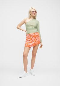 Νεαρή γυναίκα με πράσινο τοπ ενός ώμου, μικρή φούστα με πορτοκαλί λουλούδια, λευκά αθλητικά παπούτσια και ροζ γυαλιά ηλίου, στέκεται με το χέρι στη μέση.