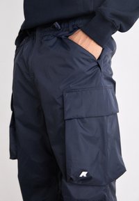 Personne portant un pantalon cargo bleu marine avec de grandes poches latérales et une main placée dans la poche avant droite.