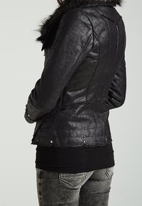 Veste noire en similicuir avec col, finition texturée, détails à franges et accents de quincaillerie argentée le long de l'ourlet. Vue arrière montrée.