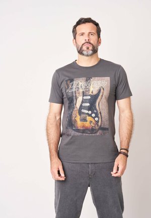 Man met donker haar en baard, dragend een donkergrijs t-shirt met een afbeelding van een vintage elektrische gitaar, staand voor een effen lichte achtergrond.