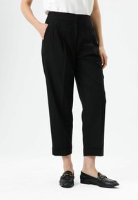 Zwarte, op maat gemaakte broek met een hoge taille, geplooide voorkant en zijzakken. Afgewerkt met omgeslagen boorden, gecombineerd met zwarte loafers.