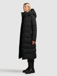 Longue doudoune noire avec capuche, dotée d'un design matelassé, d'une fermeture éclair frontale et de deux poches latérales. Associée à des bottines noires.