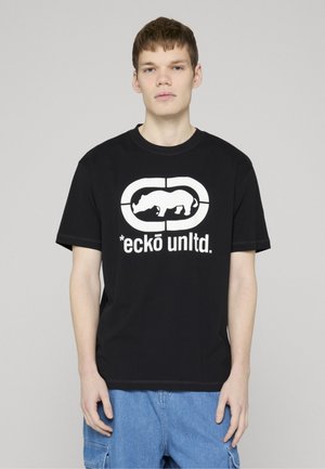 Jeune homme portant un t-shirt noir avec un logo rhinocéros blanc et le texte « eckō unltd. », assorti à un short cargo bleu, debout devant un fond gris.