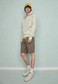Un hoodie beige avec des cordons, une chemise rayée vert olive en dessous, un short kaki et des baskets crème. Une casquette jaune complète la tenue.