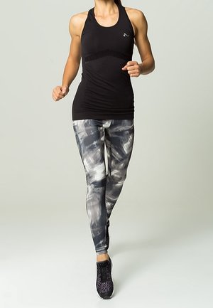 Mujer haciendo jogging con camiseta sin mangas negra y mallas grises con estampado sobre fondo liso.