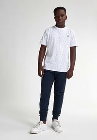 Camiseta blanca de manga corta con un sutil patrón de texto, combinada con pantalones jogger azul marino. El modelo lleva zapatillas deportivas blancas y está de pie frente a un fondo liso.