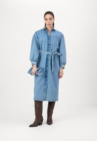 Denim srajčna obleka z dolgimi balonastimi rokavi, pasom in sprednjim žepom. Nositi jo s svetlo rjavimi škornji do kolen s peto in držati ujemajočo se torbico.