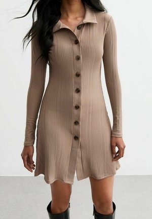 Kvinde iført en beige ribbet kjole med lange ærmer og knapper, kombineret med sorte knæhøje støvler, stående mod en ensfarvet baggrund.