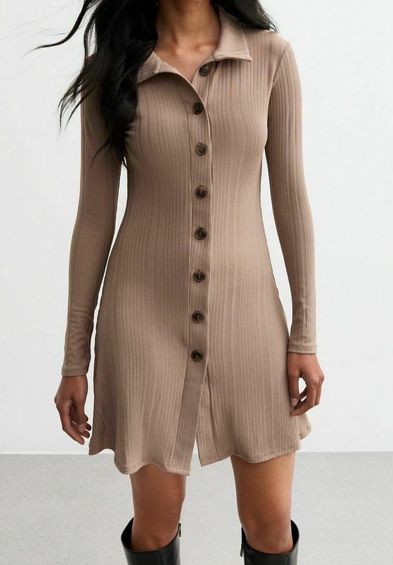 Femme portant une robe beige côtelée à manches longues boutonnée, associée à des bottes noires montant jusqu'aux genoux, debout devant un fond uni.