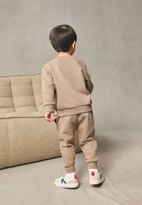 Brauner Sweatshirt- und Hosen-Set aus Baumwollmaterial, mit gerippten Bündchen, dazu beigefarbene Sneakers mit blauen und roten Akzenten. Kind von hinten gesehen.