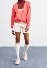 Felpa color block corallo e marrone con zip a metà, patch con logo; abbinata a pantaloni corti bianchi e calze testurizzate fino al ginocchio. Scarpe chiare.