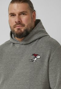Grauer Hoodie aus weichem Stoff mit einem kleinen, in Rot und Weiß gestickten Snoopy-Logo, das ein verspielt gestaltetes Charakterdesign zeigt.