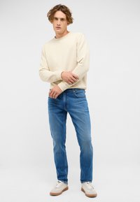 Beige sweatshirt med långa ärmar, blå jeans och krämfärgade sneakers. Enkel, avslappnad outfit med en lös passform och inga synliga mönster.