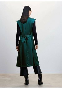 Długi zielony, satynowy kamizelka z przewiązanym w pasie, bez rękawów, zestawiona z czarnymi długimi rękawami i szerokimi czarnymi spodniami, noszona z botkami na obcasie.