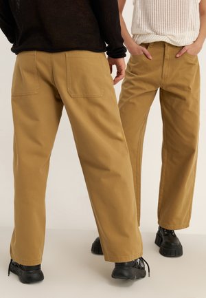 Khaki szerokie spodnie z bawełny, z dwoma tylnymi kieszeniami i solidną fakturą. Noszone z czarnymi butami na masywnej podeszwie.