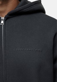 Hoodie grigio scuro con zip realizzato in tessuto morbido. Presenta una chiusura frontale a zip e il testo del marchio impresso "GOODFORNOTHING" sul petto.