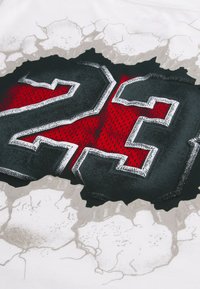 Αριθμός 23 με έντονα μαύρα γράμματα και λευκό περίγραμμα πάνω σε κόκκινο υφασμάτινο φόντο, εμφανιζόμενος σαν να σχίζει μια ραγισμένη λευκή επιφάνεια.