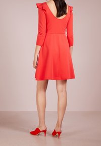 Robe rouge à manches trois-quarts, avec des détails volants sur les épaules, un dos nu et une jupe évasée. Associée à des mules rouges à bout pointu.