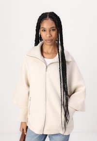 Beige lynlåsjakke med bred krave, tekstureret stof og sidelommer, parret med lyseblå jeans.