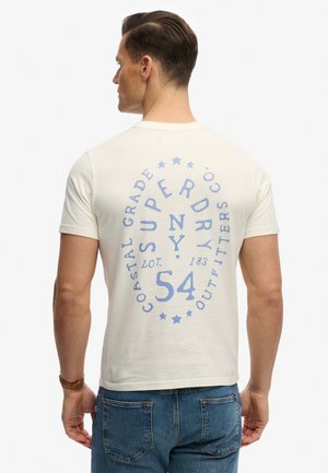 Mand iført hvid T-shirt med blå cirkulær "Superdry"-tekst og stjernedesign på ryggen, kombineret med blå jeans.