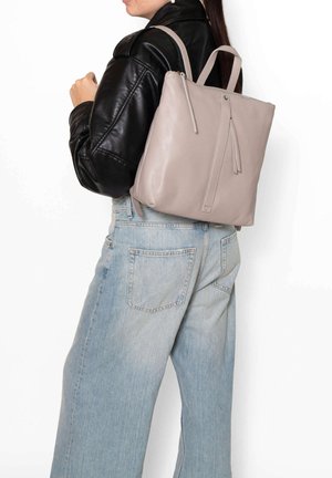 Donna con giacca di pelle nera e jeans azzurri che porta uno zaino in pelle beige chiaro con dettagli in cerniera sulla schiena.