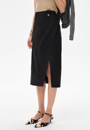 Kobieta ubrana w czarną, owijaną spódnicę z guzikami w talii, beżowy top bez rękawów, trzymająca ciemną kurtkę i szpilki w wzór krowi.