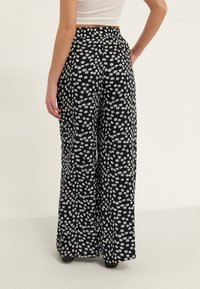 Pantalon noir avec un motif floral de marguerites blanches, coupe à jambes larges, taille élastique et texture de tissu lisse, vu de dos.