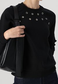 Sudadera negra con detalles de ojal redondeado, textura suave y puños acanalados. Combinada con un bolso de hombro de cuero negro.