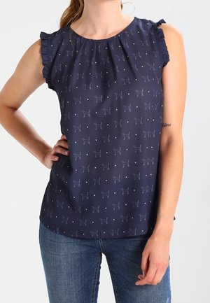 Blusa sin mangas azul marino con patrón de pequeños perros y puntos, detalles de volantes en los hombros, usada con vaqueros azules, mano en la cadera.