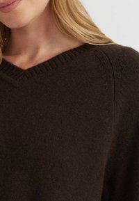 Donkerbruin gebreid sweater met een V-hals ontwerp, met ribbelafwerking en een zachte textuur. Toont detail van de schoudernaden.