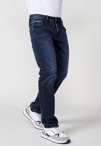 Jeans din denim albastru închis cu croi drept, având buzunare frontale, fermoar și decolorări subtile. Asortat cu adidași albi și gri.