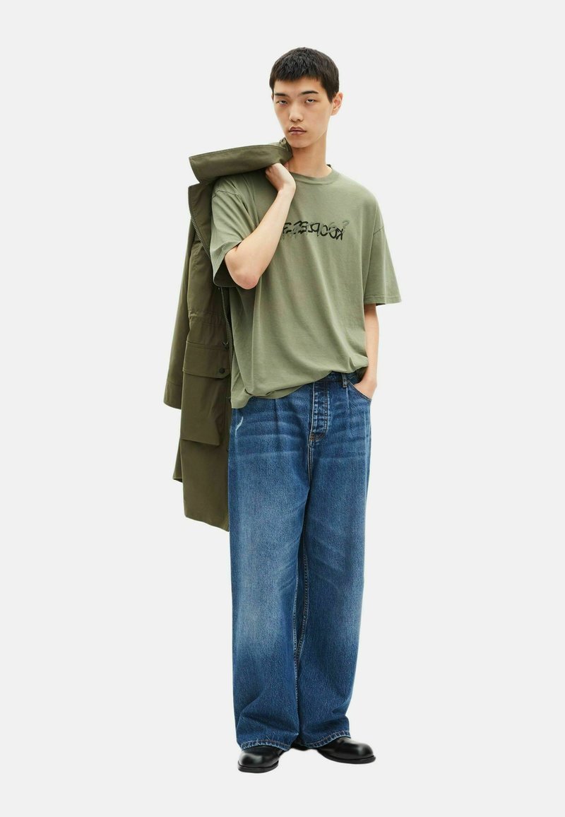 Camiseta verde oliva con estampado gráfico, combinada con vaqueros de pierna ancha azules y zapatos negros. El modelo lleva una chaqueta verde oliva sobre el hombro.