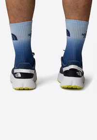 Modré a šedé tenisky s texturovaným povrchem, s bílým akcentem na patě a jasně žlutou podrážkou. V kombinaci s gradientními modrými ponožkami.