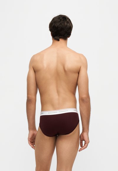 Homme debout, dos à la caméra, portant un slip rouge foncé avec une ceinture blanche et un marquage DSQUARED2 sur un fond uni.