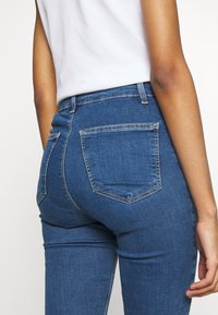 Blå denimjeans med en figursydd design, som har två bakfickor och synliga sömmar. Texturen är slät med en liten stretch.