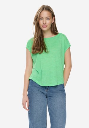 Kvinde med langt lyst brunt hår iført en løs grøn kortærmet skjorte og blå jeans, stående foran en ensfarvet hvid baggrund.