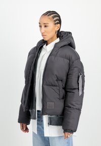 Opgeblazen zwarte jas met een capuchon, rits aan de voorkant en zakken. Heeft een label op de mouw en een gestructureerde zoom. Draag over een wit shirt.