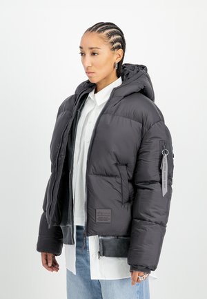 Opgeblazen zwarte jas met een capuchon, rits aan de voorkant en zakken. Heeft een label op de mouw en een gestructureerde zoom. Draag over een wit shirt.