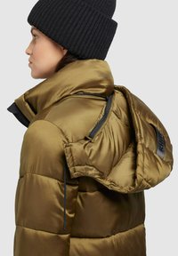Doudoune vert olive avec une finition brillante, dotée d'une capuche en fourrure, de zips et de détails de couture bien visibles. Bonnet en tricot noir porté.