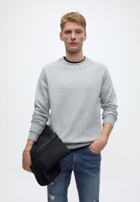 Šedá mikina s raglánovými rukávy, kulatým výstřihem a černým lemováním; opotřebované modré džíny; drží černou tašku, volný střih, minimalistický design.