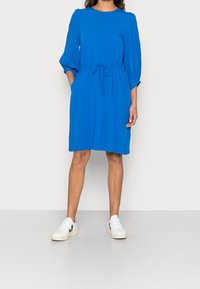 Sininen polvipituinen mekko, jossa on pyöreä pääntie, ryppyiset puff-hihat, säädettävä vyötärönauha ja sileä kangas. Yhdistetty valkoisiin tennareihin.