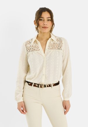 Mujer de cabello oscuro vestida con blusa de encaje color crema con cuello y cinturón estampado, de pie frente a un fondo blanco liso.