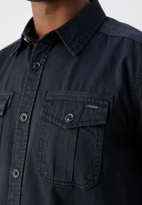 Dunkelblaufarbenes Button-Up-Hemd aus Baumwolle mit zwei Brusttaschen mit Klappen, einem Kragen und Metallknöpfen. Strukturierter Stoff mit dezentem Glanz.