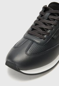 Zwarte sportieve schoen van glad en geperforeerd leer. Voorzien van platte zwarte veters en een witte tussenzool. Gebogen neusvorm.