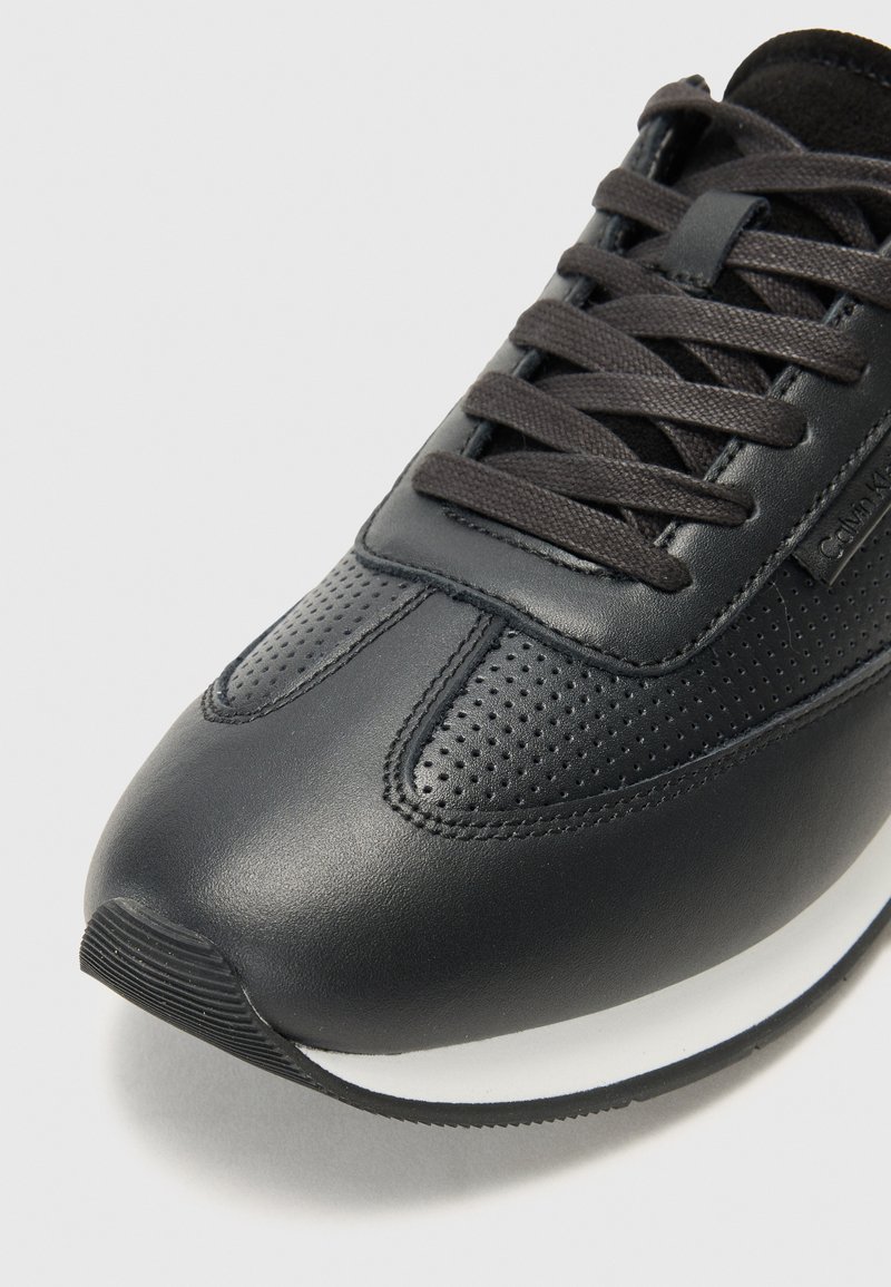 Chaussure de sport noire en cuir lisse et perforé. Elle possède des lacets noirs plats et une semelle intermédiaire blanche. Forme de bout incurvée.