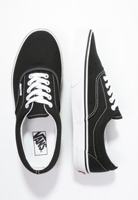 Baskets en toile noire avec lacets blancs, semelle en caoutchouc blanche, et bandes latérales noires. Présentent un logo Vans à l'intérieur et une étiquette rouge sur le talon.
