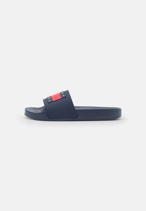 Granatowy klapek z szerokim paskiem ozdobionym czerwono-biało-czarnym prostokątnym logo oraz napisem "JEANS" wytłoczonym na boku podeszwy.