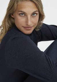 Top in tessuto blu scuro strutturato con una leggera lucentezza. Primo piano di una persona con occhi chiari, che indossa un design aderente con maniche lunghe.
