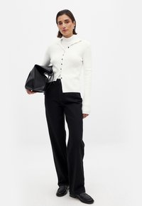 Cárdigan blanco de canalé con botones, combinado con pantalones de pierna ancha negros y mocasines negros, sosteniendo un bolso negro. Diseño minimalista, textura suave.